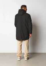 Man wearing a black coat and beige pants against a white wall