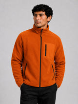 A man wearing a bright orange fleece jacket with a black front zipper, black vertical zip pockets, and a black chest pocket, paired with black pants, against a solid gray studio background.