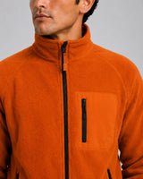 A close-up of a man wearing a bright orange fleece jacket with a stand-up collar, black front zipper, and a vertical zippered chest pocket.