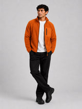 A man wearing a bright orange fleece zip-up jacket over a white t-shirt, black pants, and black boots, standing against a light gray studio background.