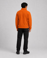 A man viewed from behind wearing a bright orange fleece pullover, black trousers, and black boots, standing against a light gray studio background.