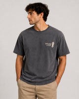 A man wearing a dark gray washed t-shirt with a small white graphic on the left chest, paired with khaki pants, standing against a light gray studio background.