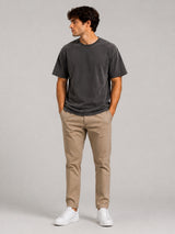 A man wearing a black washed t-shirt, beige chinos, and white sneakers, standing against a solid light gray studio background.