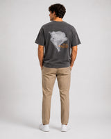 A man viewed from behind wearing a black graphic t-shirt, khaki pants, and white sneakers against a light gray studio background.