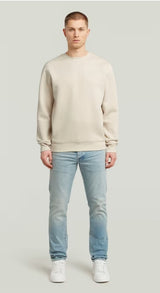 Man wearing a beige sweater and light blue jeans on a plain background