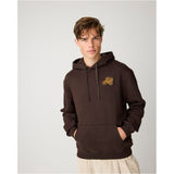 A man wearing a dark brown pullover hoodie with a small gold embroidered carriage logo on the left chest, paired with light beige trousers, set against a plain light gray studio background.