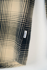 Close-up of a plaid fabric garment in beige, black, and white, showing a small white brand tag that reads 'LAKOR' against a plain white background.