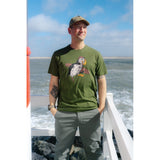 A man wearing an olive green t-shirt with a puffin graphic, standing on a white railing with the ocean and sky in the background.