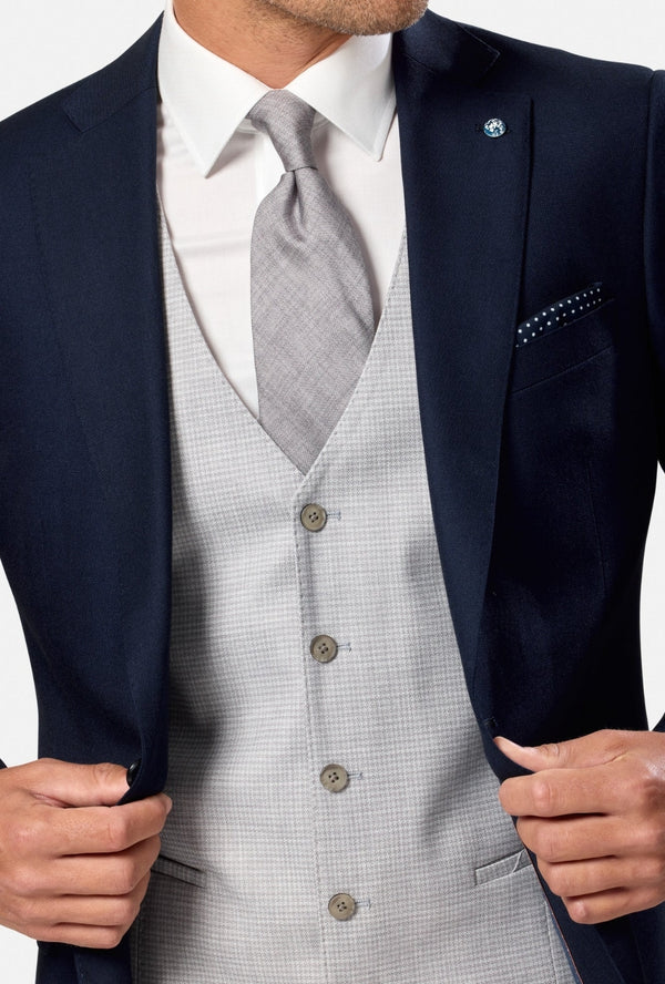 Man wearing a navy blazer, white shirt, and gray vest on a plain background