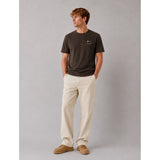 A man wearing a dark brown crew-neck t-shirt with a small smiley face graphic on the left chest, paired with beige trousers and tan suede shoes, standing against a solid gray studio background.