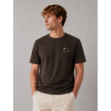 A man wearing a dark brown crewneck t-shirt with a small white smiley face graphic on the left chest, paired with light beige pants, set against a plain gray studio background.