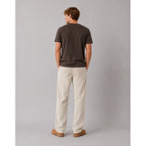 A man standing with his back to the camera, wearing a dark brown t-shirt, off-white linen trousers, and brown leather shoes against a solid gray studio background.