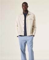 Man wearing a beige jacket over a blue shirt and light blue pants on a plain background