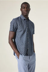 Man wearing a patterned short-sleeve shirt and light gray pants against a plain background