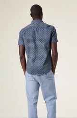 Man wearing a blue patterned shirt and light blue pants on a beige background