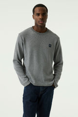 A man wearing a navy textured crewneck sweater with a small navy embroidered logo on the left chest, paired with dark blue jeans, against a plain light background.