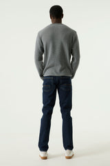 A man viewed from behind wearing a navy long-sleeve knit sweater, dark blue jeans, and white sneakers, standing against a plain light gray studio background.