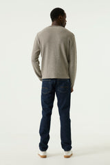 A man viewed from behind wearing a light gray knit sweater and dark blue jeans, standing against a plain white studio background.