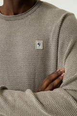 Close-up of a person wearing a textured knit crewneck sweater in taupe-beige, with a small square chest patch logo on the left chest.