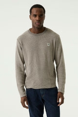 A man wearing a textured, long-sleeve crewneck sweater in a light taupe-beige color with a small square logo patch on the left chest, paired with dark blue jeans, against a plain light gray background.