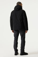 A man viewed from behind wearing a black hooded jacket, dark gray jeans, and black shoes, standing against a plain light gray studio background.