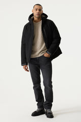A man wearing a black hooded jacket over a beige knit sweater, dark gray jeans, and black sneakers, standing against a plain light gray studio background.