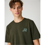 A man wearing an olive green crewneck t-shirt with a small light blue embroidered graphic on the left chest, photographed against a neutral gray background.