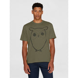 A man wearing an olive green crewneck t-shirt with a dark gray line art owl graphic on the front, paired with dark blue pants, set against a plain light gray background.