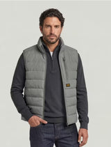 A man wearing a light gray quilted puffer vest over a dark gray quarter-zip sweater and dark blue jeans, standing against a light gray studio background.